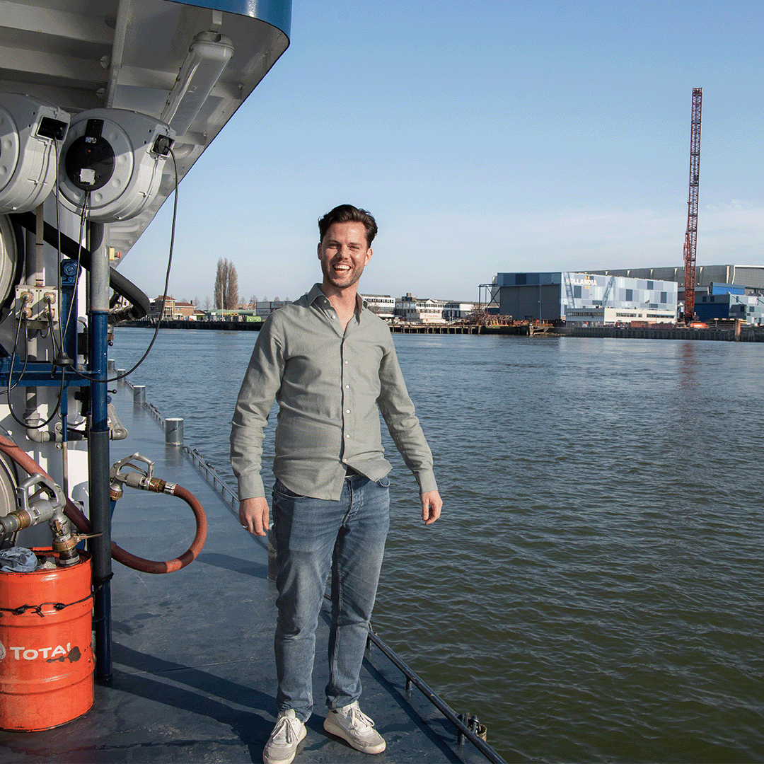 Smiling man in a grey shirt and jeans standing on the deck of a ship, with an industrial harbor and water in the background under a blue sky.